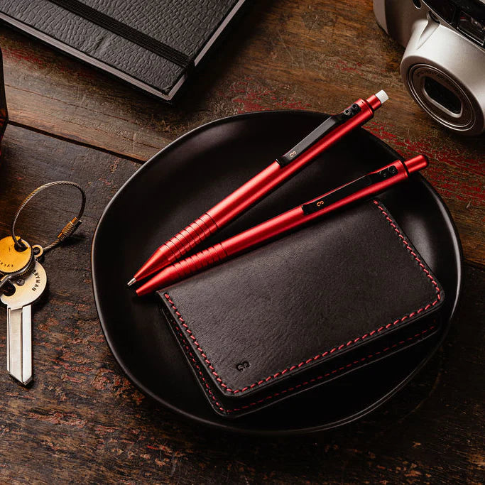 A collection of men's everyday carry items including a leather wallet, pen, multitool, and keychain arranged on a textured surface.
