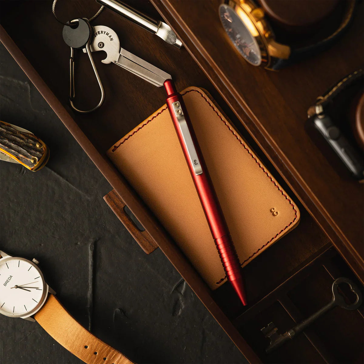 A modern, stylish workspace featuring a minimalist desk setup with a sleek pen and pencil set, a leather wallet, multitool, keychain, and a watch in earthy tones and metallic accents.