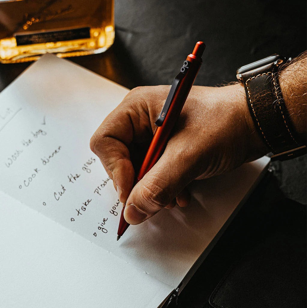 A sleek desk setup with a hand holding a Grafton Pen, writing in a notebook, embodying a modern, masculine aesthetic desk.