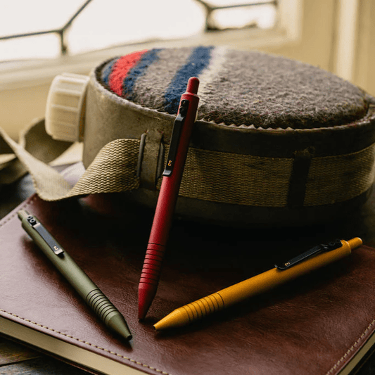 Three Grafton Pens on a brown book against a water container. 