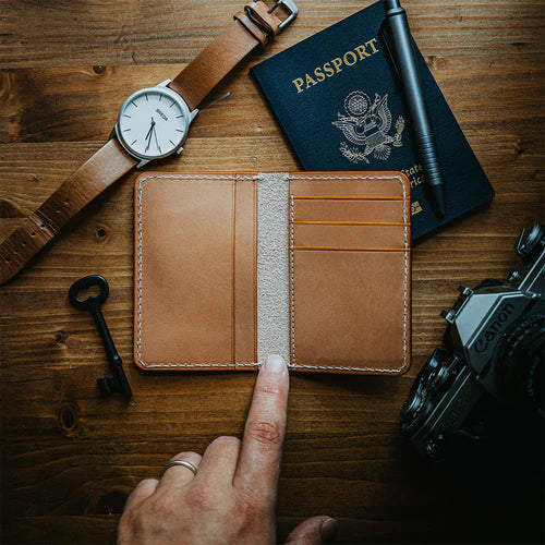 A sleek, slim bifold leather wallet on a wooden surface surrounded by a minimalist pen, multitool, stylish keychain, watch, and the edge of a camera.