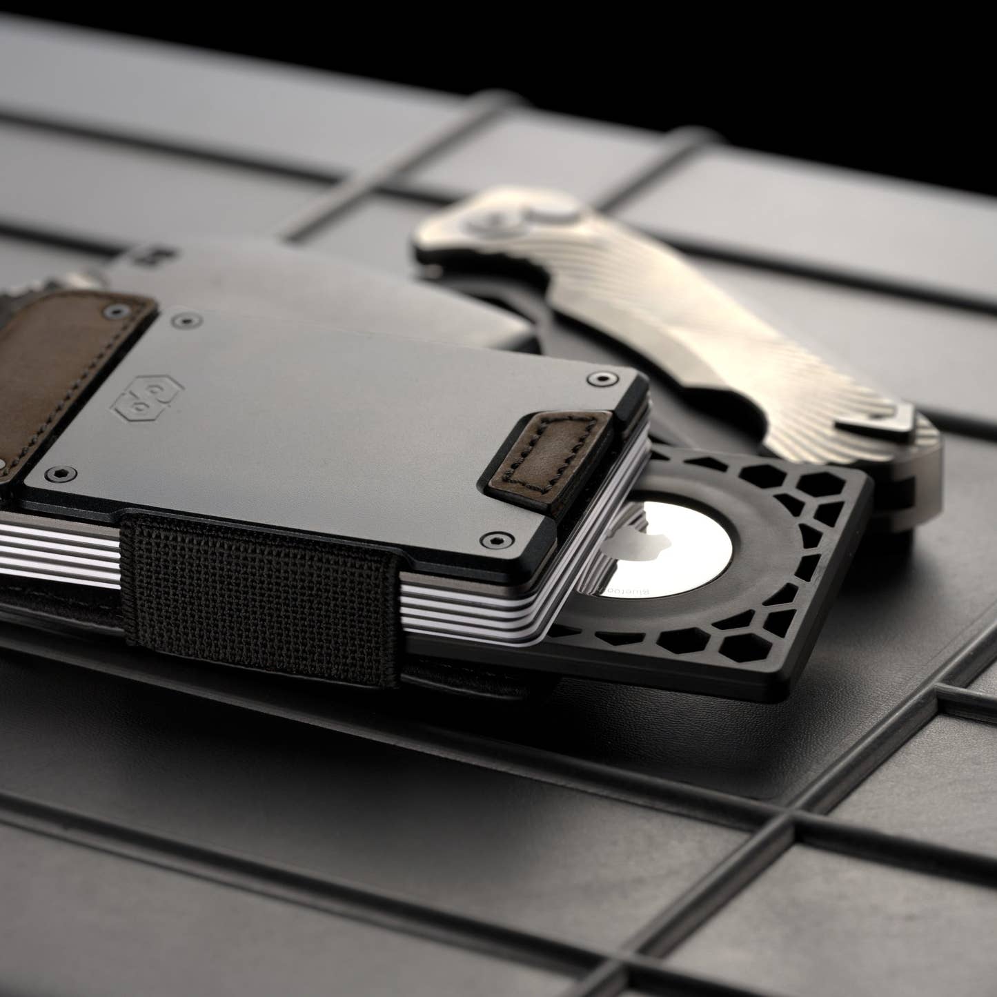 PROOF AirTag holder fitted to a gunmetal and brown leather PROOF wallet on a dark metal grate, with a folding knife alongside