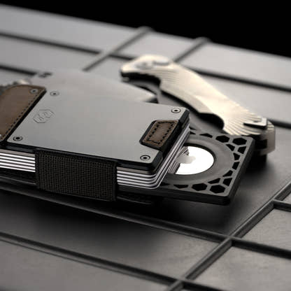 PROOF AirTag holder fitted to a gunmetal and brown leather PROOF wallet on a dark metal grate, with a folding knife alongside