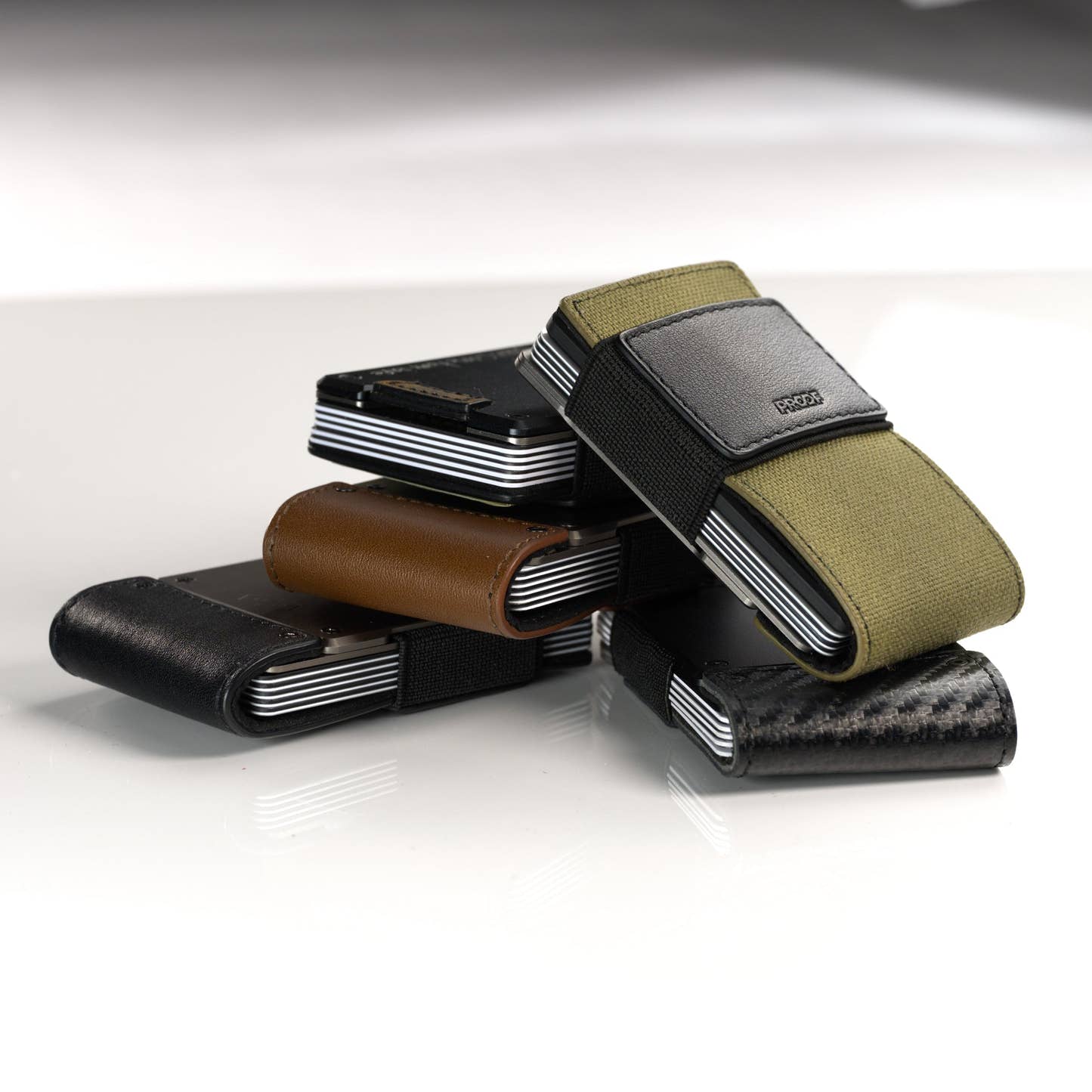 A stacked group of PROOF wallets in olive nylon, brown leather, black leather and carbon fiber; the AirTag holder itself is not shown in this frame