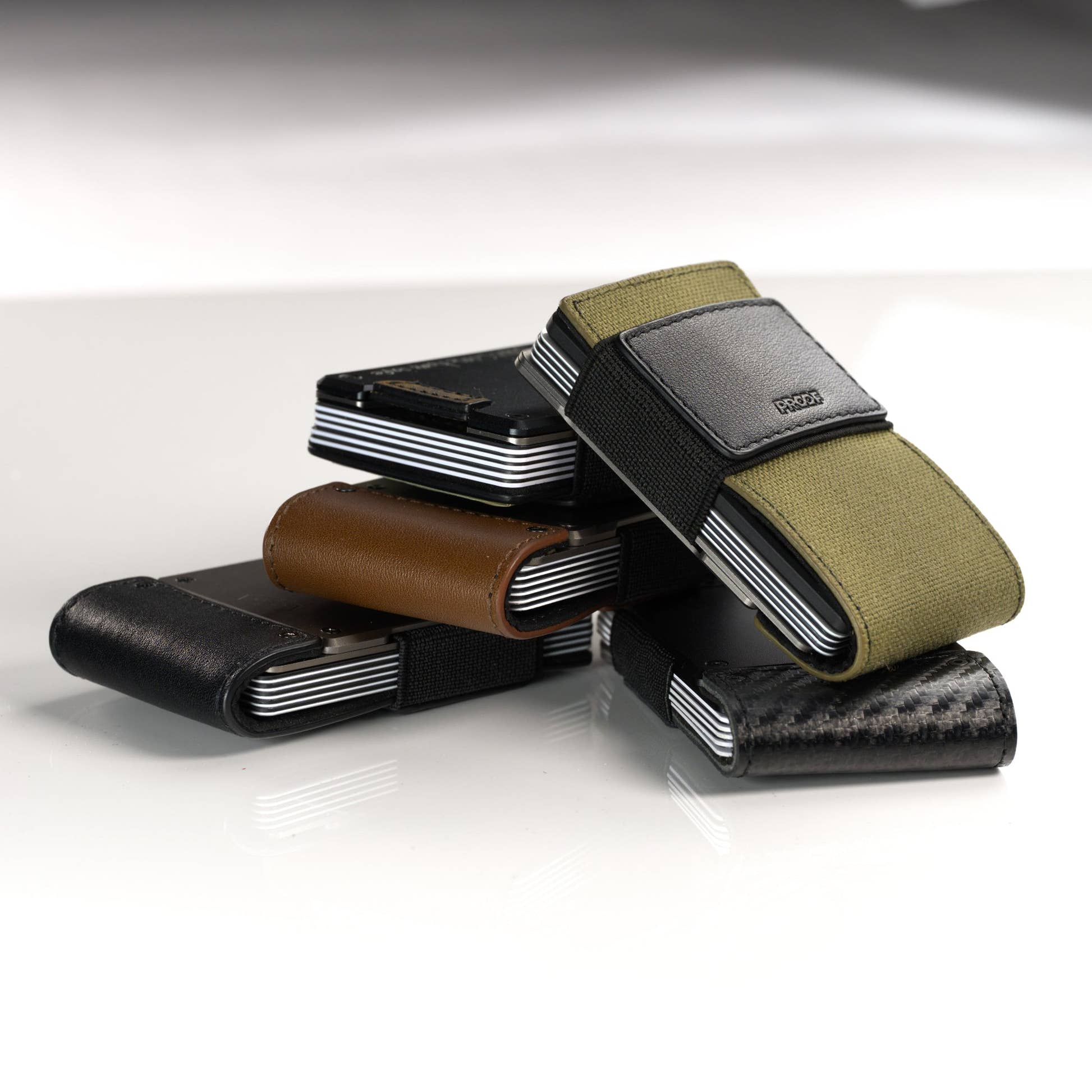 A stacked group of PROOF wallets in olive nylon, brown leather, black leather and carbon fiber; the AirTag holder itself is not shown in this frame