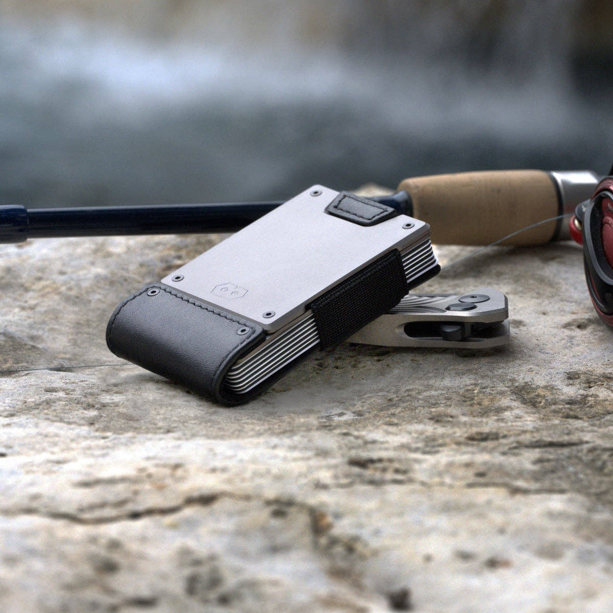 PROOF Apex wallet on a river rock beside a fishing rod and pliers, showing stacked cards inside the aluminum frame