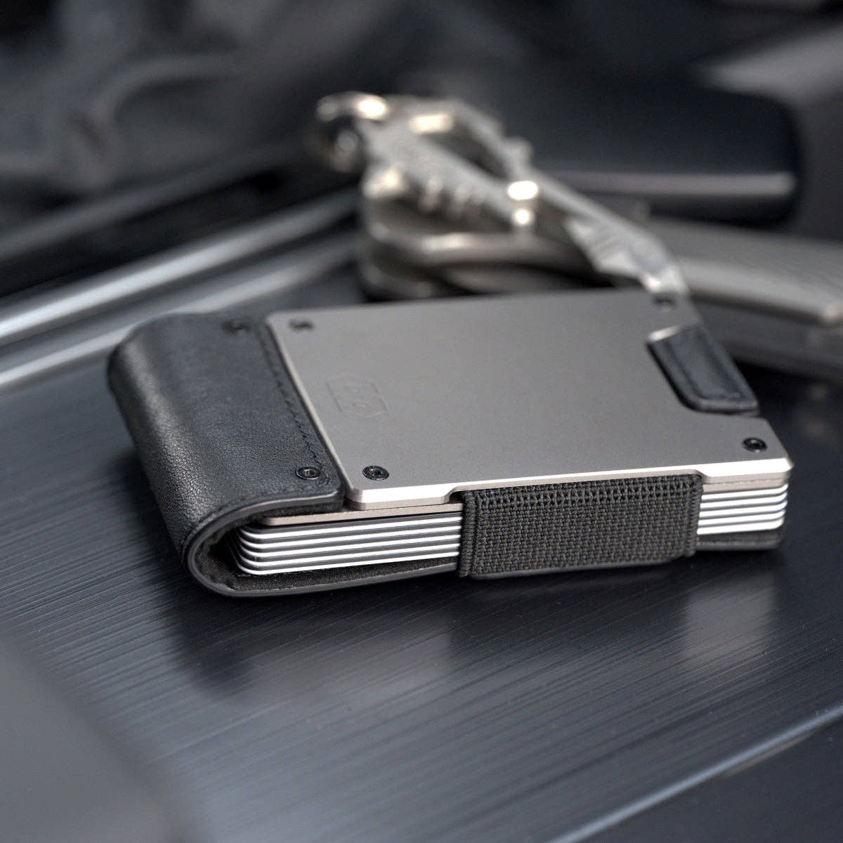 PROOF Apex wallet in matte black leather and satin gunmetal on a dark brushed console beside a set of keys