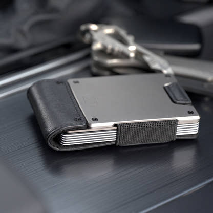 PROOF Apex wallet in matte black leather and satin gunmetal on a dark brushed console beside a set of keys