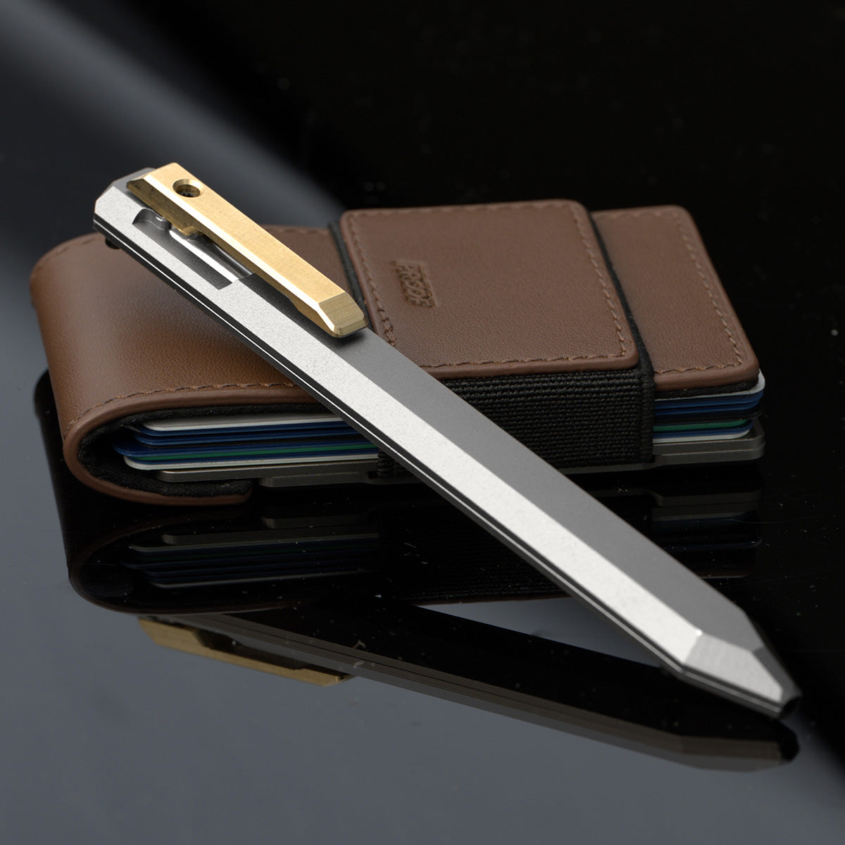 PROOF Vanguard pen leaning against a brown leather PROOF wallet on a dark reflective surface