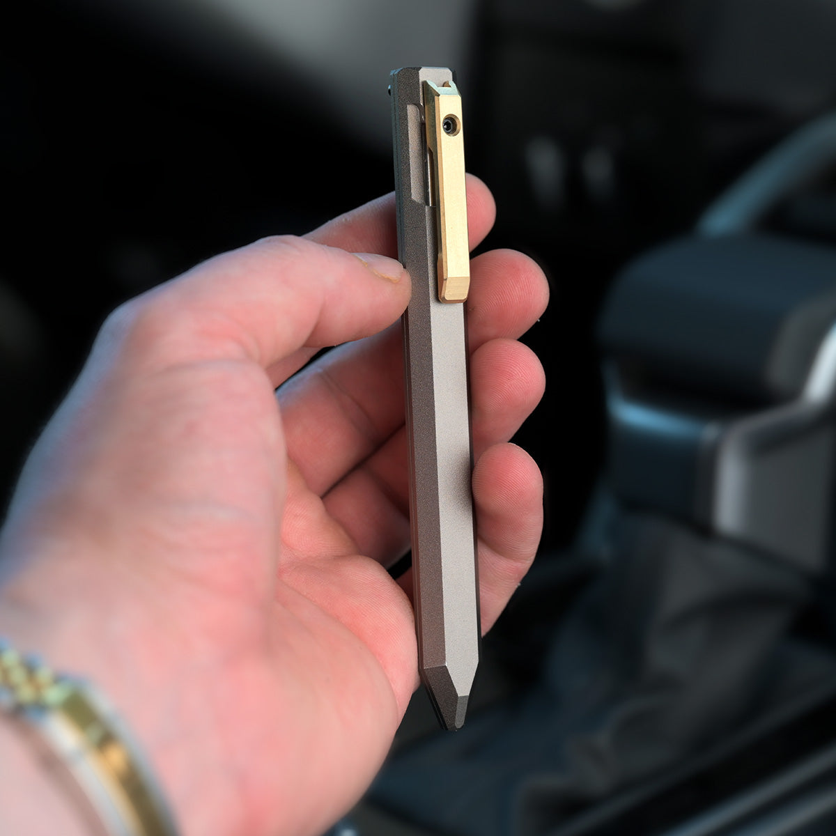 Hand holding the PROOF Vanguard pen upright inside a vehicle, brass clip and bolt facing the camera