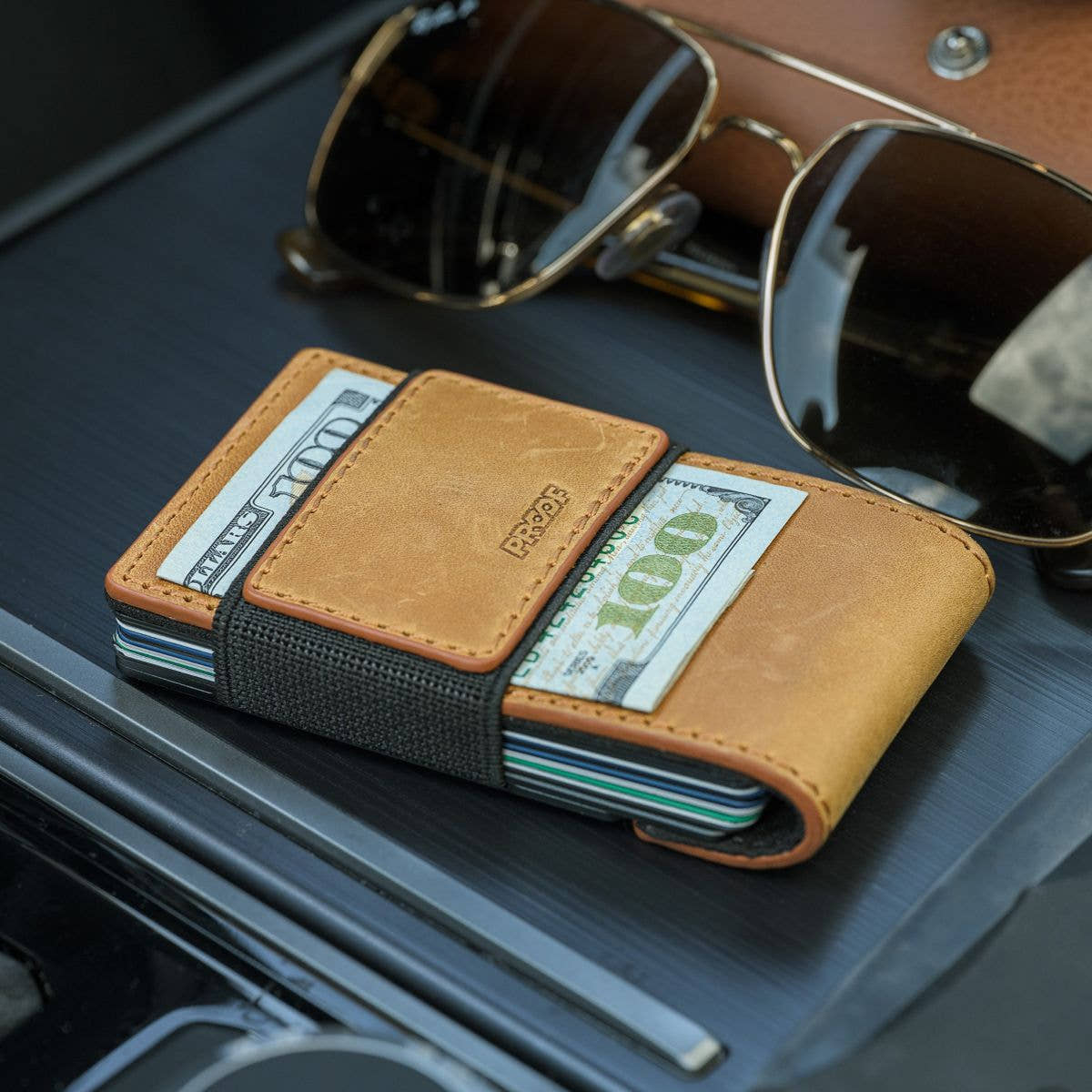 Tobacco leather PROOF Executive wallet loaded with cards and cash under its elastic strap, beside aviator sunglasses