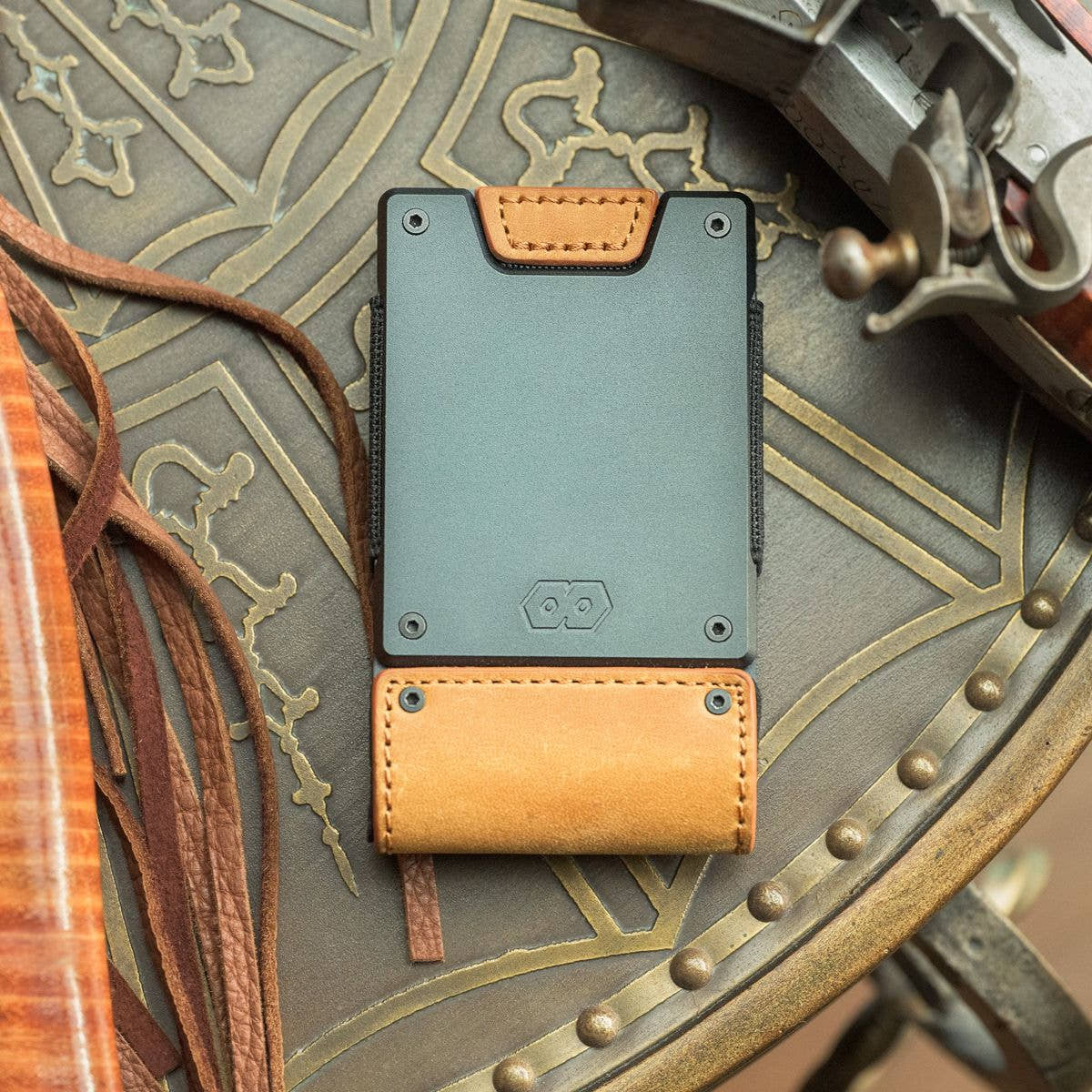 Top-down view of the PROOF Executive wallet on a studded leather surface beside antique brass hardware, gunmetal plate and tobacco leather strap facing up