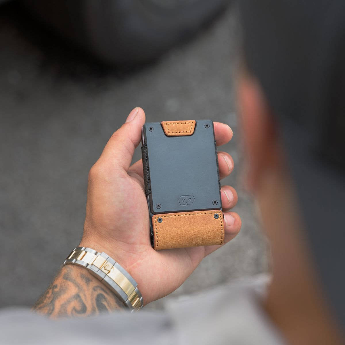 Man's hand holding the PROOF Executive wallet, dark gunmetal front plate with embossed hexagon logo above the tobacco leather strap