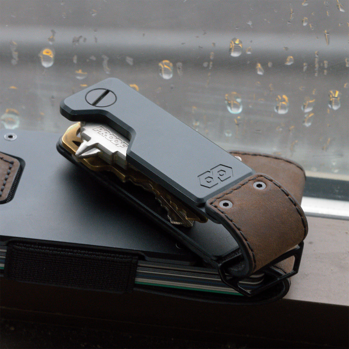 PROOF Founder key holder on top of a PROOF wallet against a rain-streaked window, a brass key swung partway out