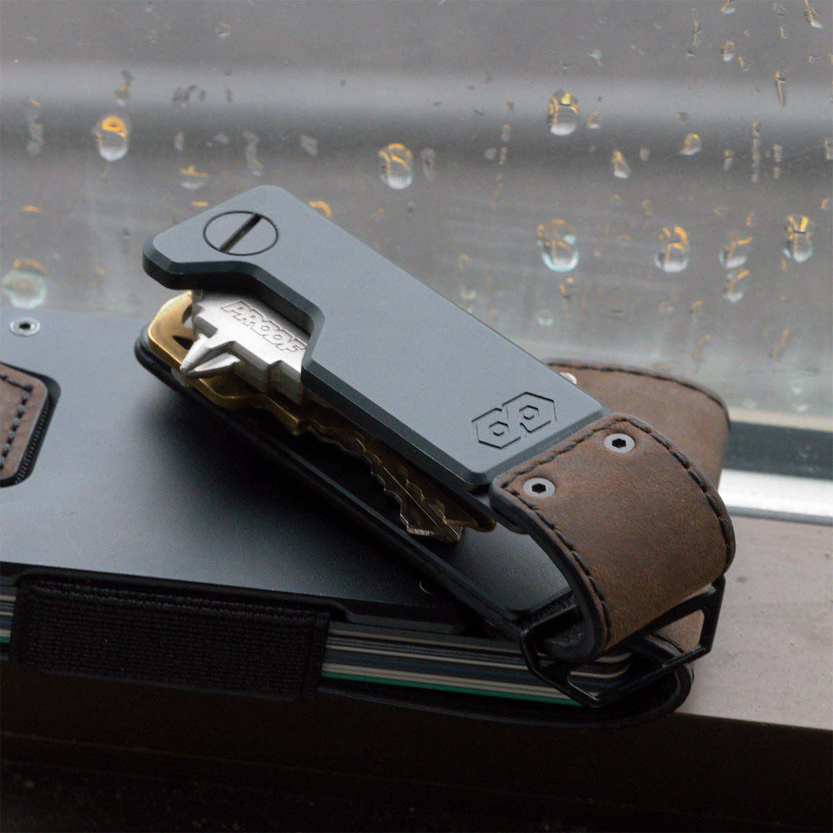 PROOF Founder key holder on top of a PROOF wallet against a rain-streaked window, a brass key swung partway out
