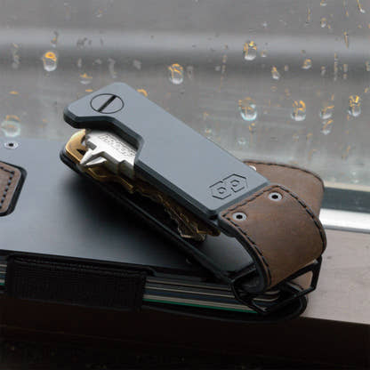 PROOF Founder key holder on top of a PROOF wallet against a rain-streaked window, a brass key swung partway out