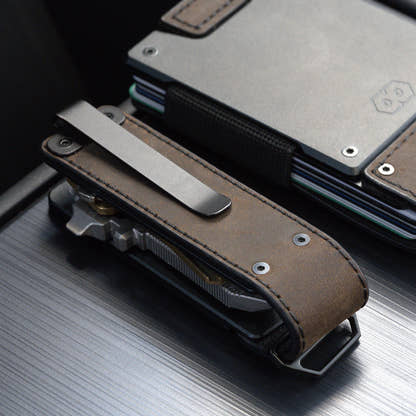 Back of the PROOF Founder key holder on dark wood, steel pocket clip over the brown leather panel, leaning against a PROOF wallet