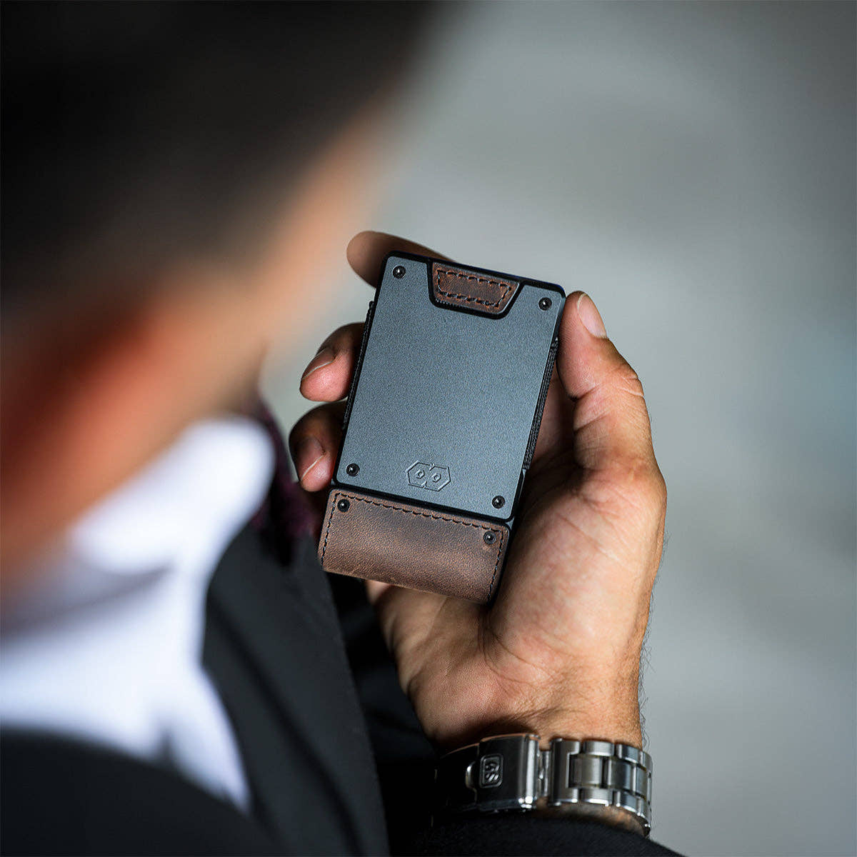 Man in a suit and steel watch holding up the PROOF Founder wallet, dark gunmetal front plate with embossed hexagon logo above the bomber leather strap