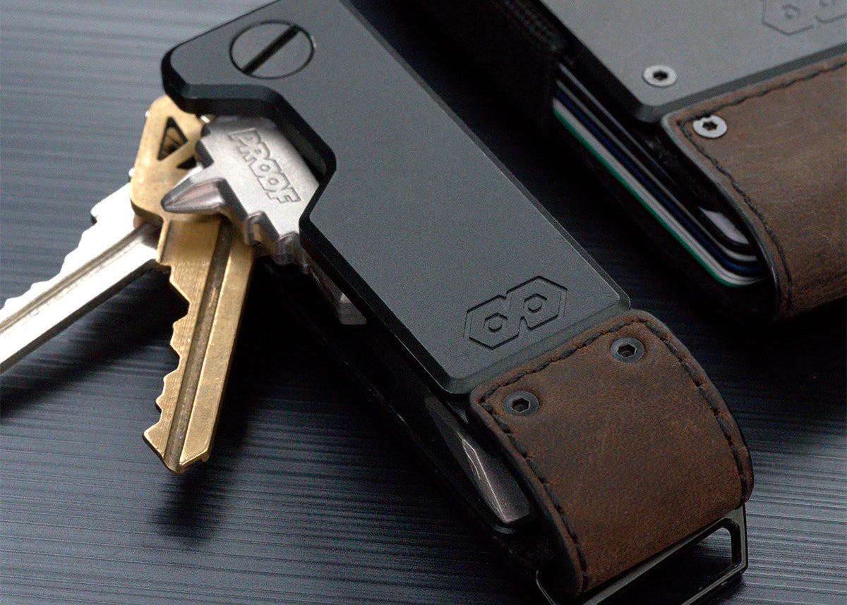 Close-up of the PROOF Founder key holder with brass and steel keys fanned open, brown leather strap and embossed hexagon logo in focus