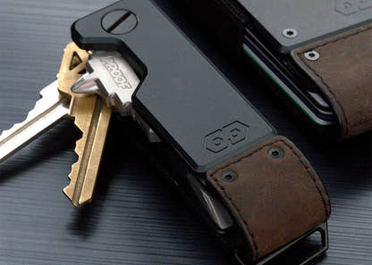 Close-up of the PROOF Founder key holder with brass and steel keys fanned open, brown leather strap and embossed hexagon logo in focus