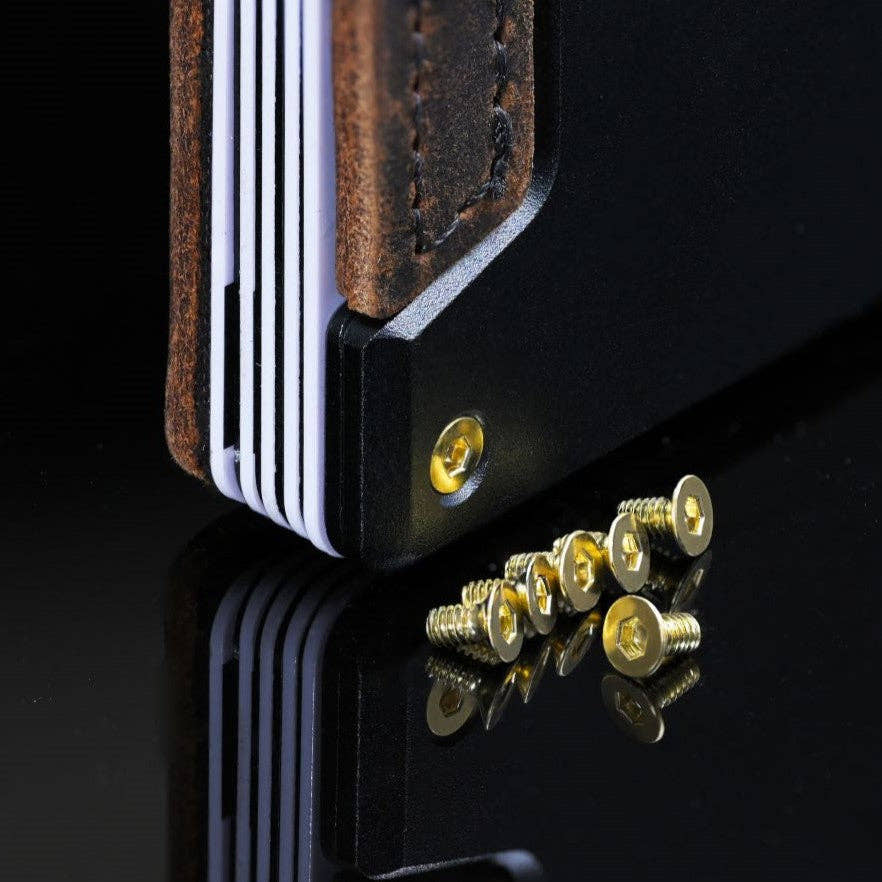 Gold PROOF wallet bolts scattered on a dark reflective surface next to a wallet corner fitted with a matching gold bolt