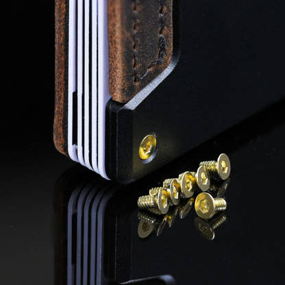 Gold PROOF wallet bolts scattered on a dark reflective surface next to a wallet corner fitted with a matching gold bolt