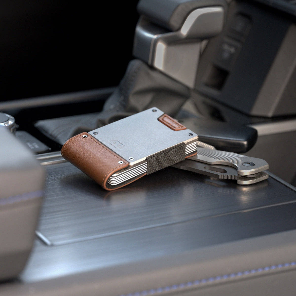 PROOF Legacy wallet on a vehicle centre console beside a set of keys, satin gunmetal plate above the tan leather strap