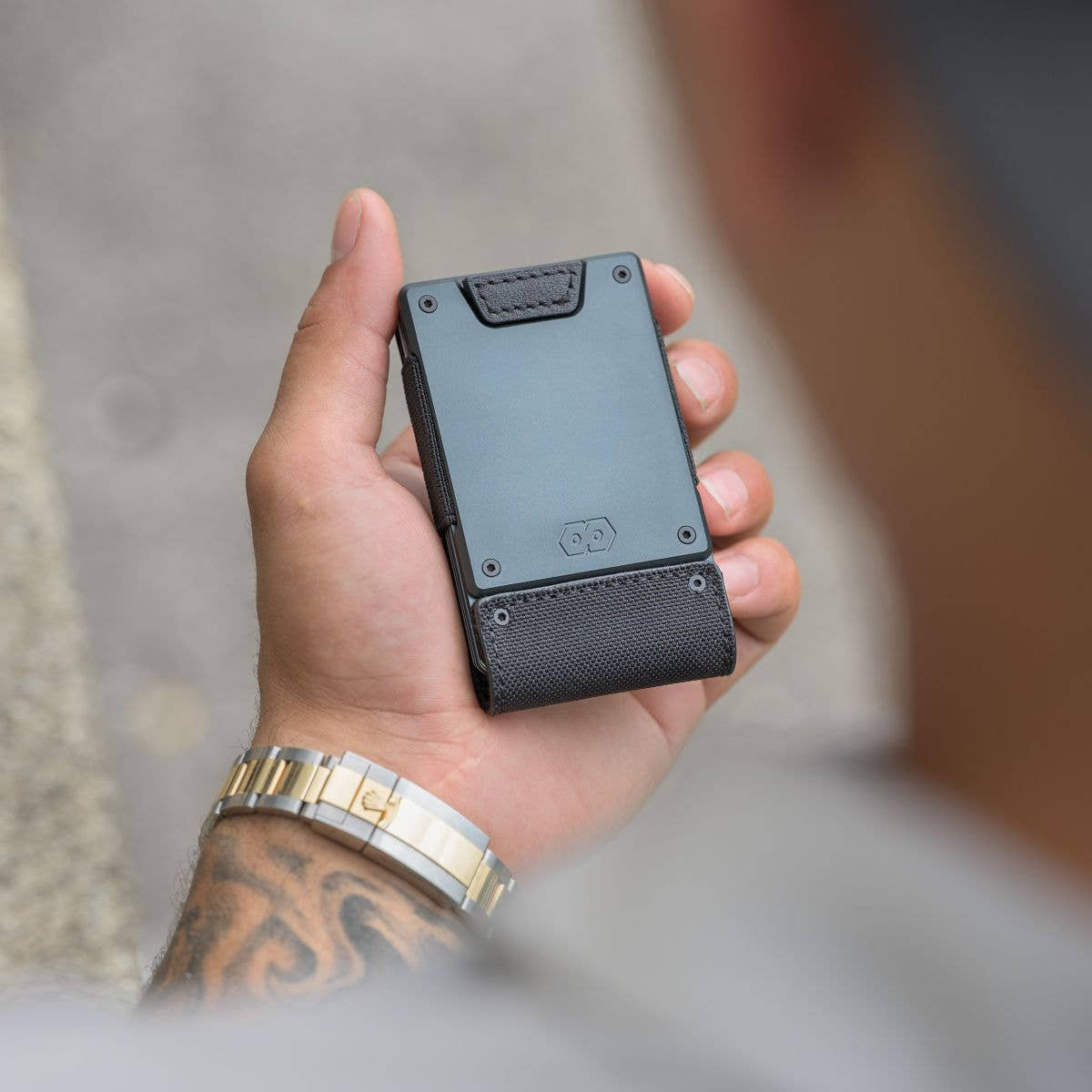 Hand holding the PROOF Monolith wallet, dark gunmetal plate above the black ballistic nylon strap