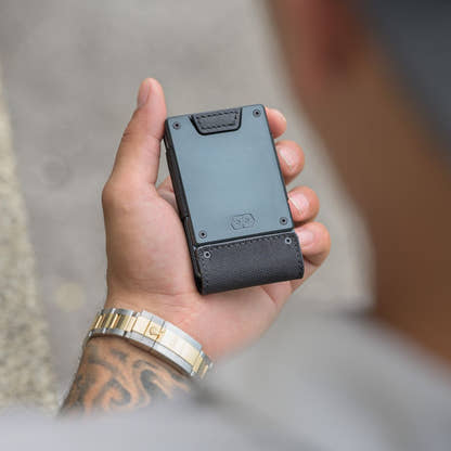 Hand holding the PROOF Monolith wallet, dark gunmetal plate above the black ballistic nylon strap