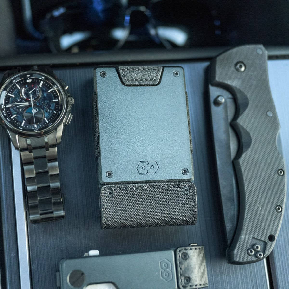PROOF Monolith wallet on a dark console beside a chronograph watch, folding knife and phone