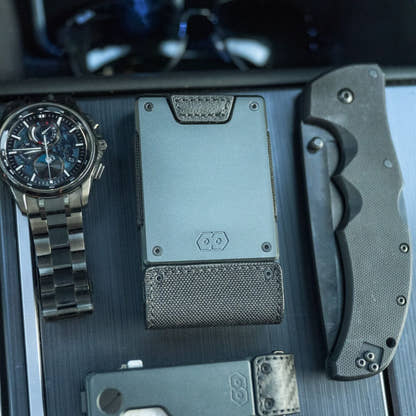 PROOF Monolith wallet on a dark console beside a chronograph watch, folding knife and phone