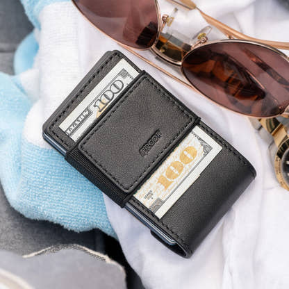Black full-grain leather back of the PROOF Obsidian wallet with folded bills under the elastic strap, resting on a towel beside rose-gold aviator sunglasses
