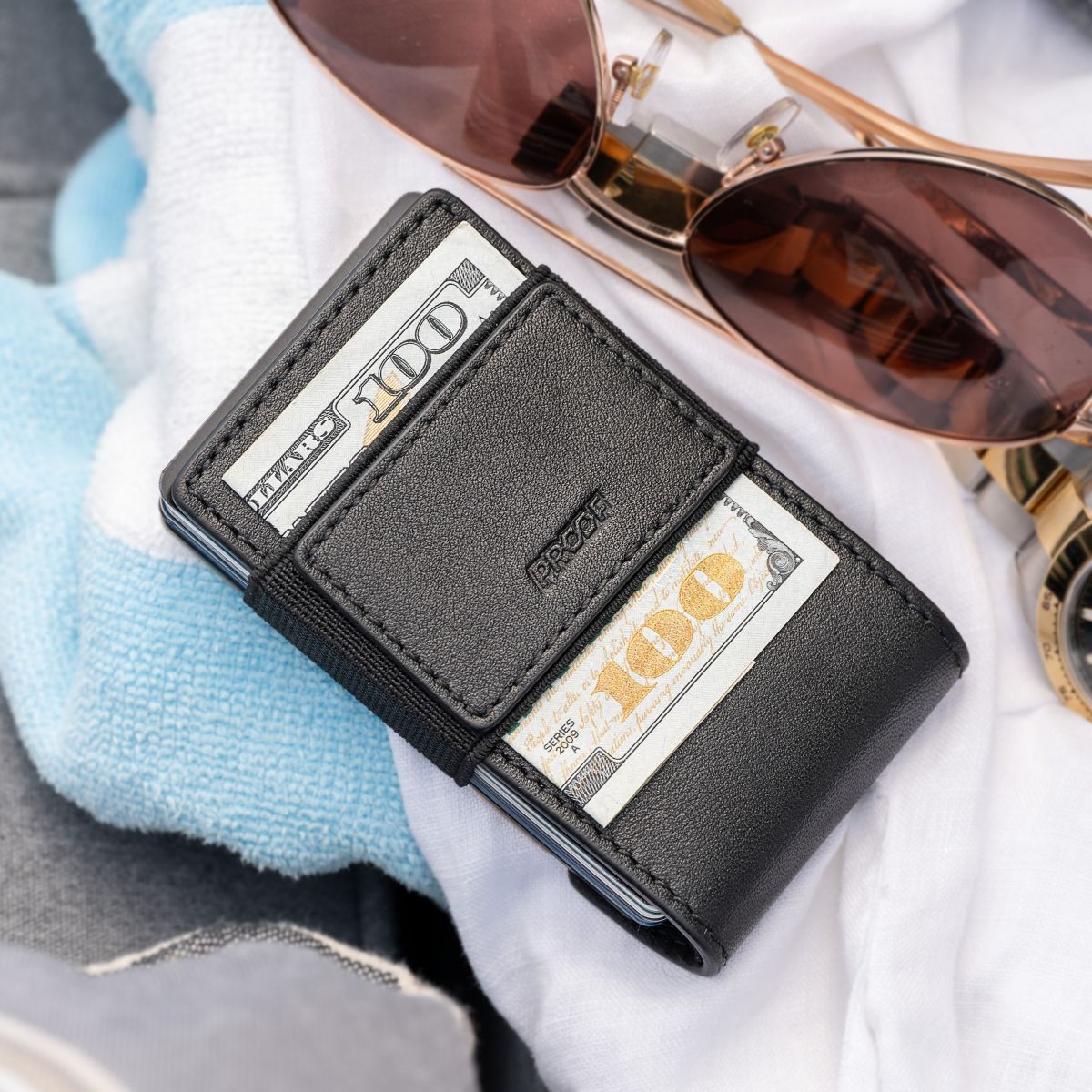 Black full-grain leather back of the PROOF Obsidian wallet with folded bills under the elastic strap, resting on a towel beside rose-gold aviator sunglasses