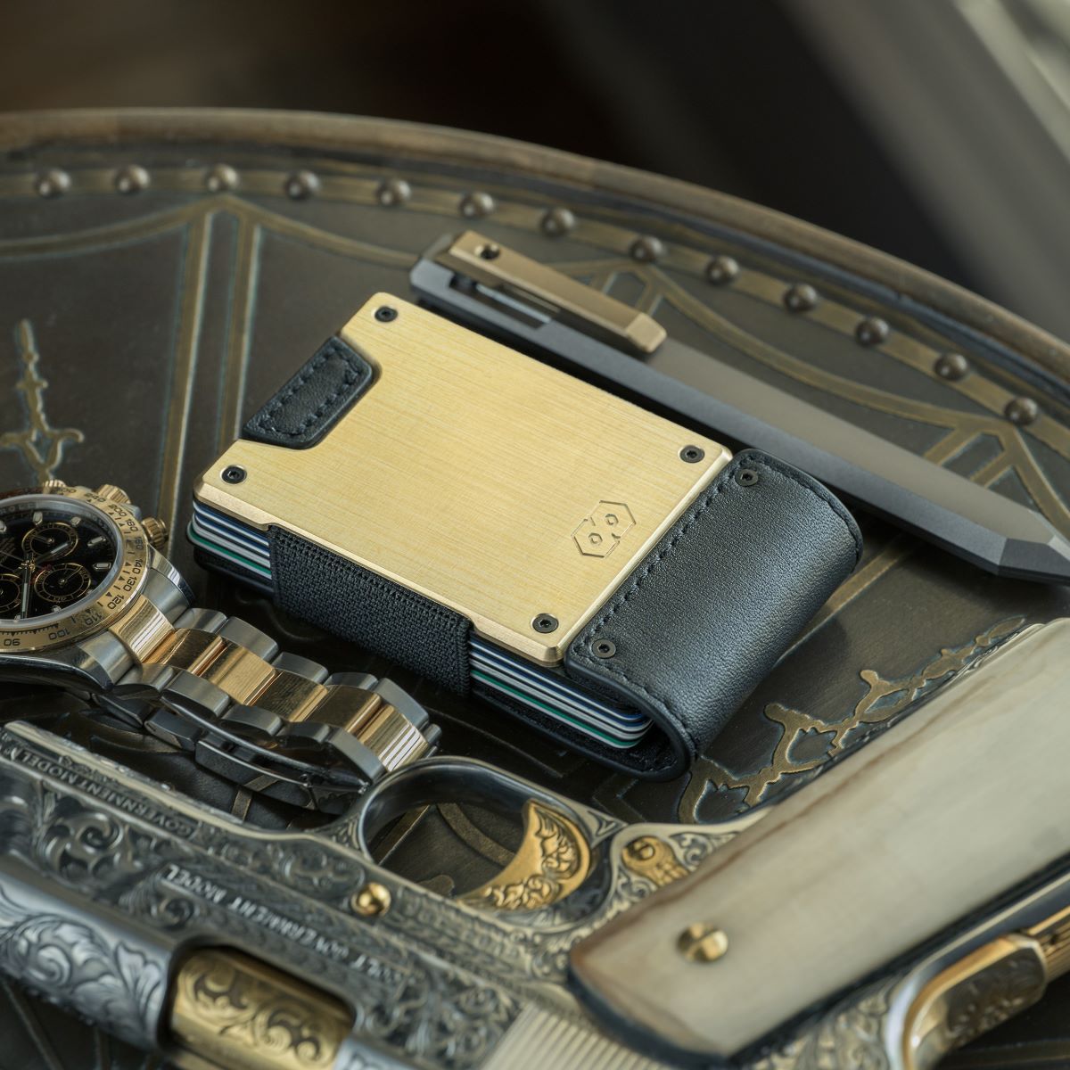 PROOF Obsidian wallet resting on an engraved metal tray beside a two-tone wristwatch and a folding knife, brushed brass plate catching the light