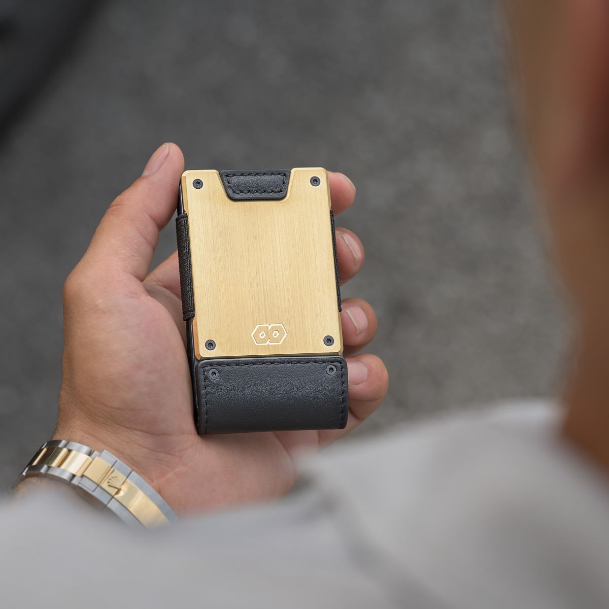 Hand holding the PROOF Obsidian wallet, brushed brass face with embossed hexagon logo above a black leather corner tab