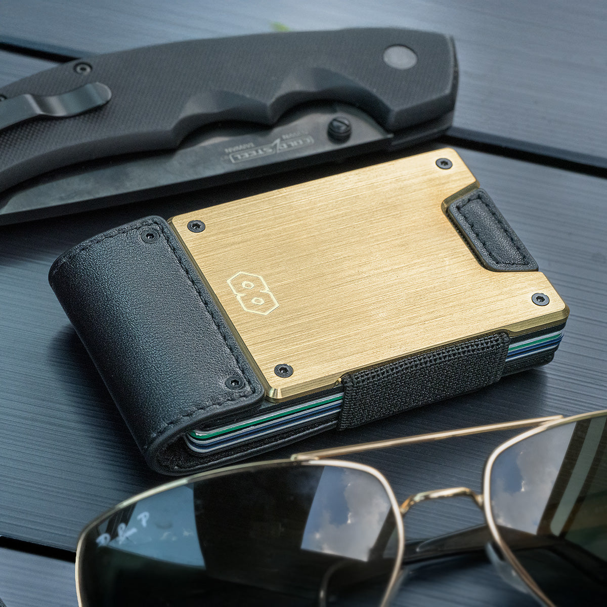 PROOF Obsidian wallet on a dark dashboard beside a black folding knife, brushed brass plate and stacked card edges in view