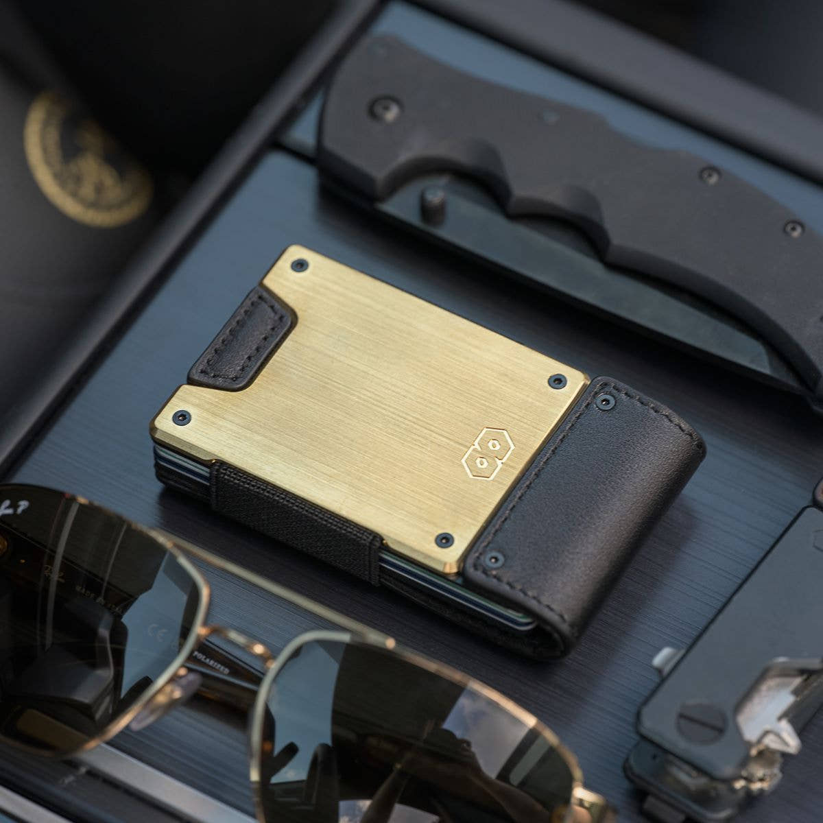 PROOF Obsidian wallet on a dark slatted dash with a folding knife and gold-framed aviator sunglasses, brushed brass plate facing up