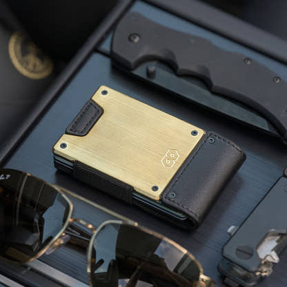 PROOF Obsidian wallet on a dark slatted dash with a folding knife and gold-framed aviator sunglasses, brushed brass plate facing up