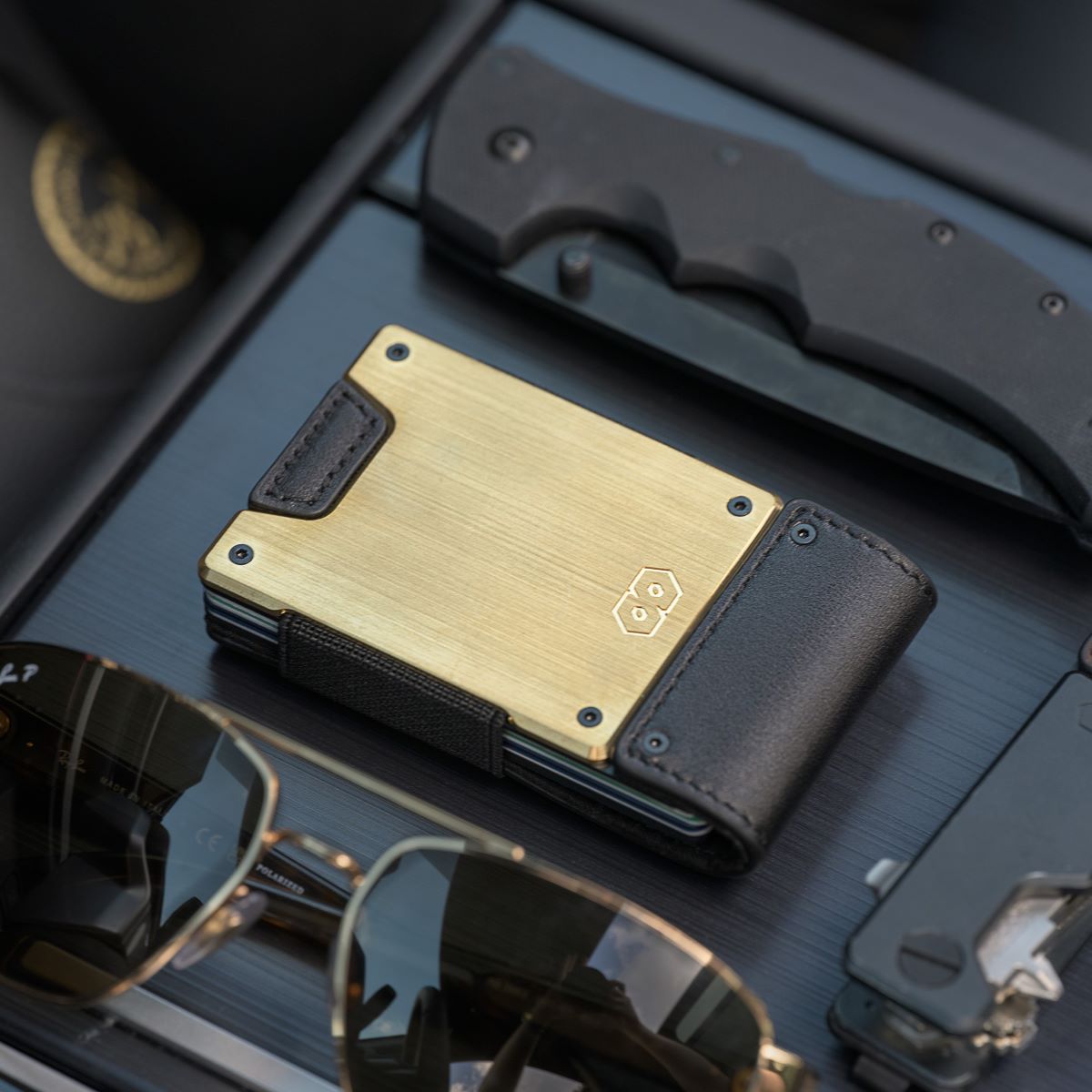 PROOF Obsidian wallet on a dark slatted dash with a folding knife and gold-framed aviator sunglasses, brushed brass plate facing up
