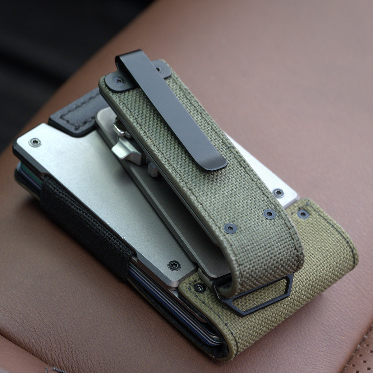 Back of the PROOF Operator key holder on a tan leather car seat, black pocket clip mounted over the olive drab nylon panel