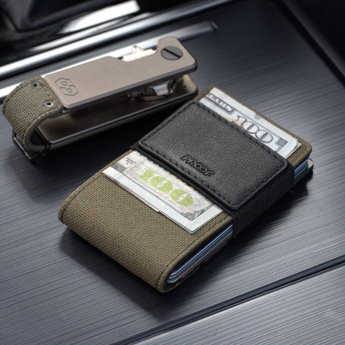 PROOF Operator wallet holding hundred-dollar bills under its black leather strap, with the matching Operator key holder behind it on a dark ribbed surface
