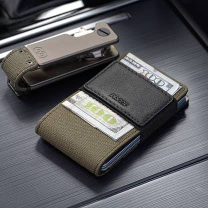 PROOF Operator wallet holding hundred-dollar bills under its black leather strap, with the matching Operator key holder behind it on a dark ribbed surface