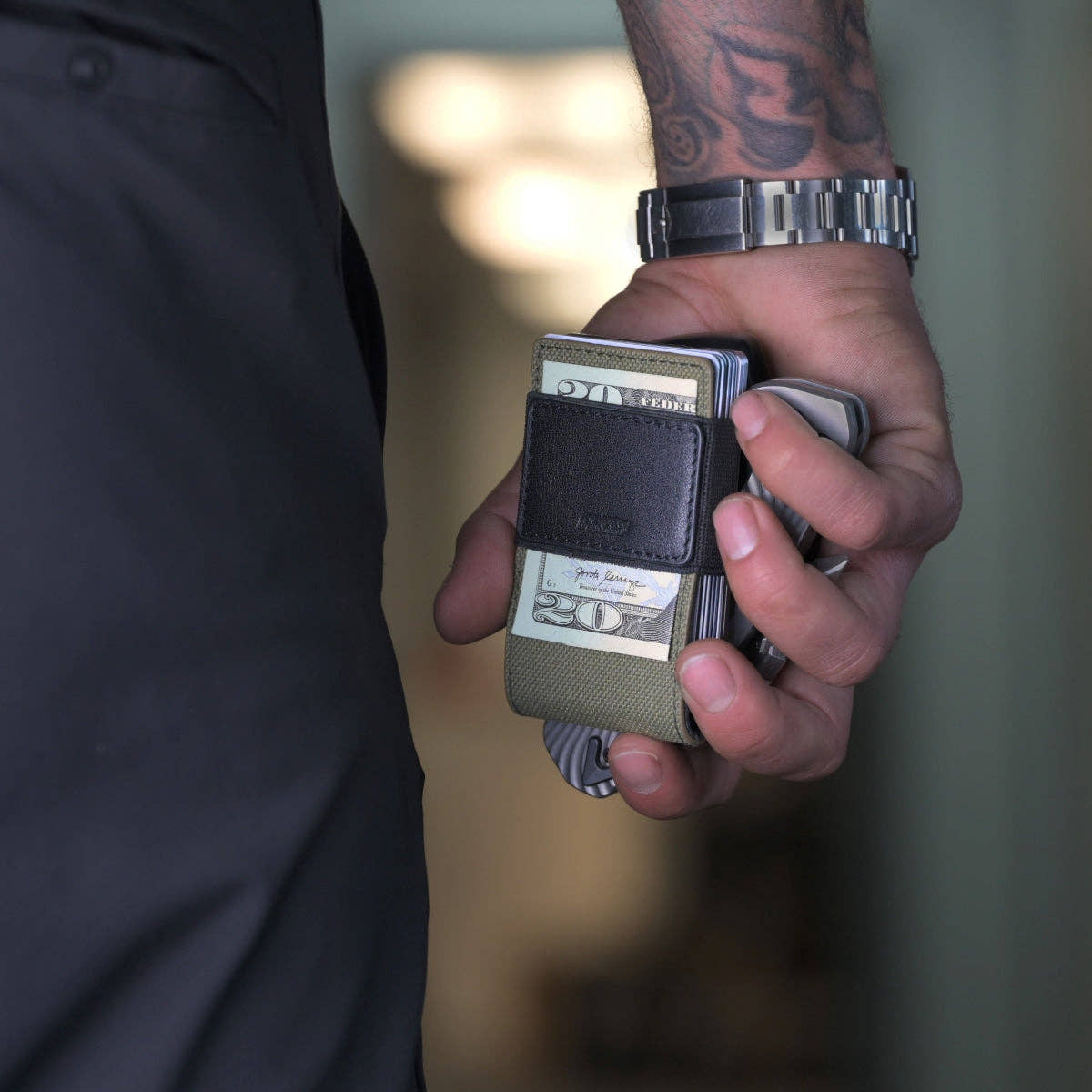 Man holding the PROOF Operator wallet with cash and cards gripped under the strap, wristwatch and tattooed forearm in frame