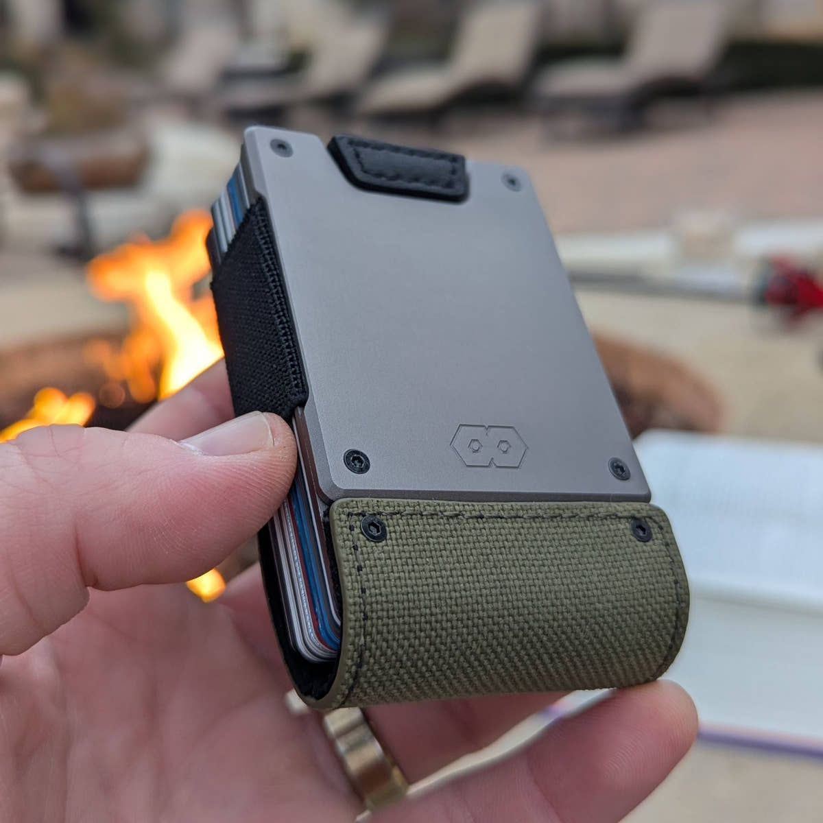 Hand holding the PROOF Operator wallet in front of an outdoor fire, satin gunmetal plate over the olive drab nylon strap