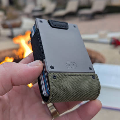 Hand holding the PROOF Operator wallet in front of an outdoor fire, satin gunmetal plate over the olive drab nylon strap