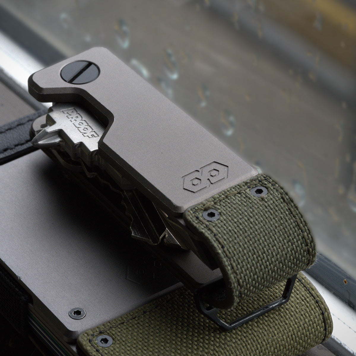 PROOF Operator key holder resting on a PROOF wallet against a rain-streaked window, olive drab nylon strap and steel loop visible