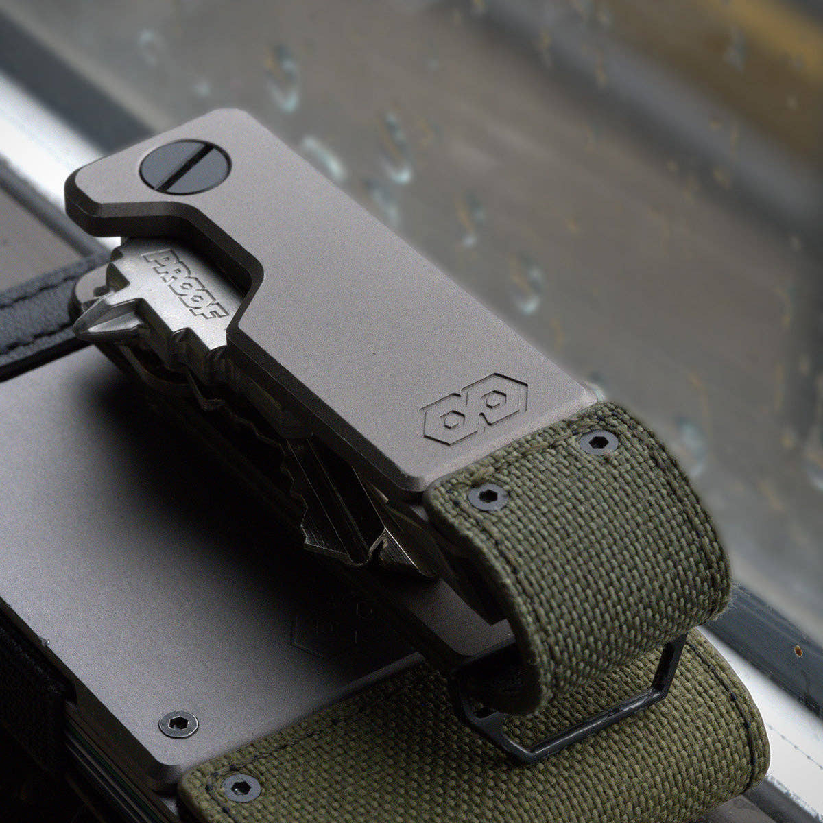 PROOF Operator key holder resting on a PROOF wallet against a rain-streaked window, olive drab nylon strap and steel loop visible