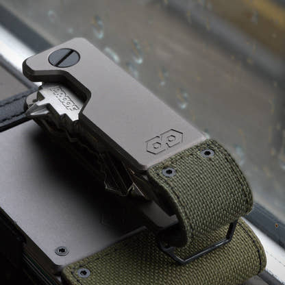 PROOF Operator key holder resting on a PROOF wallet against a rain-streaked window, olive drab nylon strap and steel loop visible