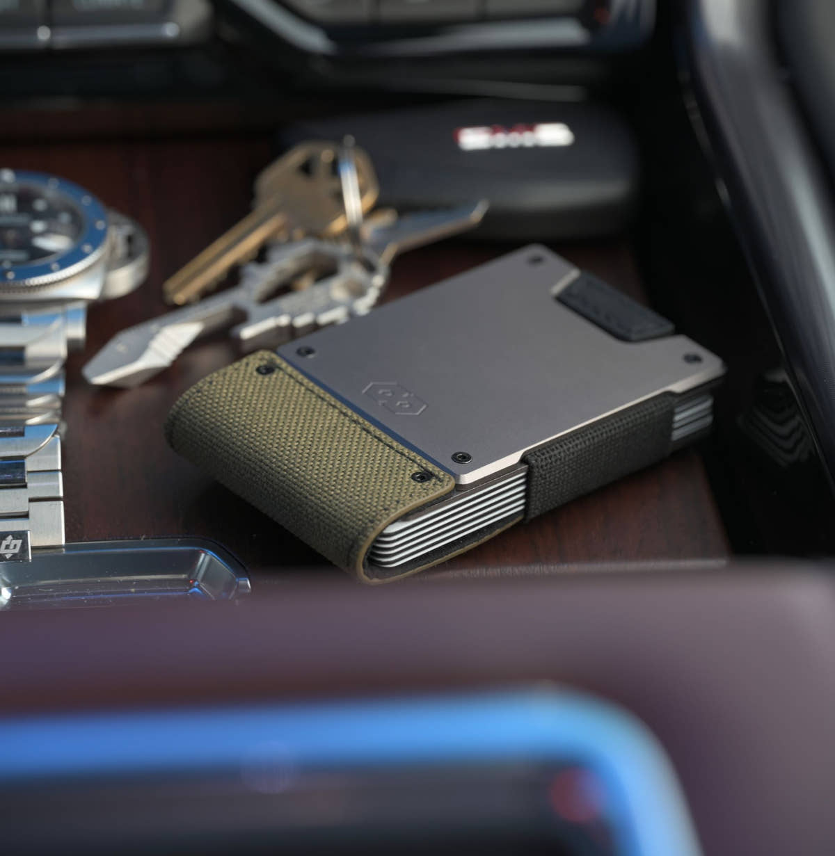PROOF Operator wallet on a wood-trimmed console with a dive watch and keys, olive drab nylon strap facing the camera