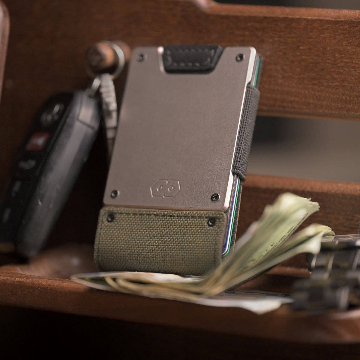 PROOF Operator wallet standing on a wooden surface beside a car key fob and loose cash, satin gunmetal plate above olive drab nylon